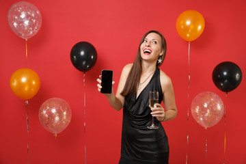 Cheerful young woman in black dress holding glass of champagne, mobile phone with blank black empty screen on bright red background air balloons. Happy New Year, birthday mockup holiday party concept.