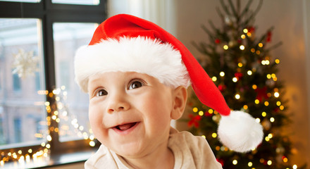 childhood, holidays and people concept - close up of happy little baby boy in santa hat over christmas tree lights background