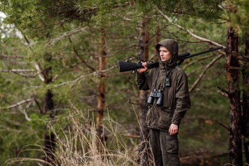 male hunter with binoculars ready to hunt, holding gun and walking in forest.