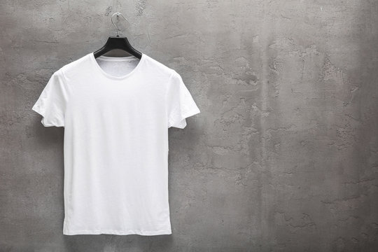 Front side of male white cotton t-shirt on a hanger and a concrete wall in the background. T-shirt without print and copyspace for your text on right side
