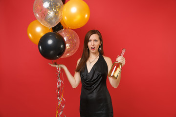 Irritated young woman in black dress spread hands celebrating hold bottle of champagne, air balloons isolated on red background. Valentine's Day, Happy New Year, birthday mockup holiday party concept.