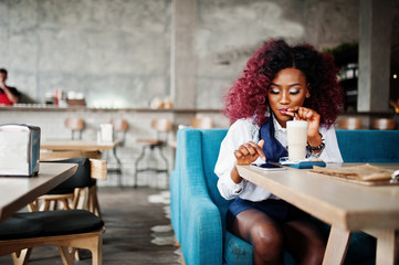 Attractive african american curly girl sitting at cafe drinking latte and looking at mobile phone.