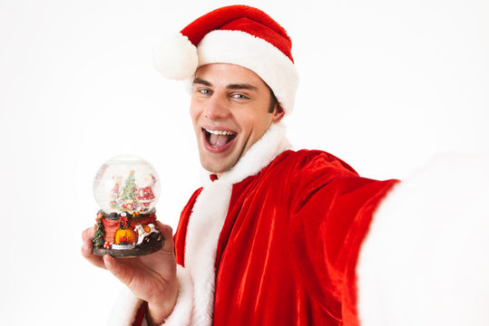 Image of european man 30s in santa claus costume and red hat holding christmas ball, isolated on white background in studio