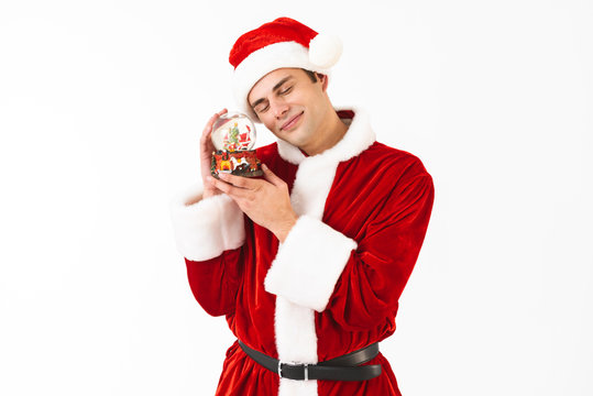 Image of attractive man 30s in santa claus costume and red hat holding christmas ball, isolated on white background in studio