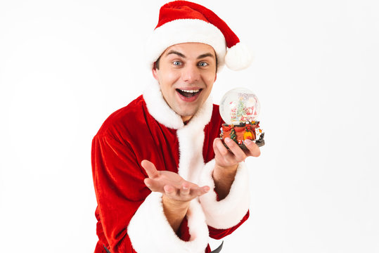 Image of handsome man 30s in santa claus costume and red hat holding christmas ball, isolated on white background in studio