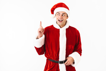 Portrait of excited man 30s in santa claus costume and red hat gesturing finger upward, isolated over white background in studio