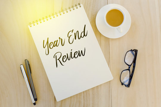 Top view of pen,sunglasses,a cup of coffee and notebook written with Year End Review on wooden background.