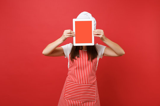 Housewife female chef cook or baker in striped apron white t-shirt toque chefs hat isolated on red wall background. Woman hold tablet pc blank screen for promotional content Mock up copy space concept