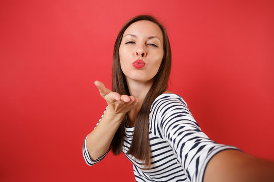 Close up selfie shot of pretty young woman in casual striped clothes blowing kisses send air kiss isolated on bright red wall background. People sincere emotions lifestyle concept. Mock up copy space.