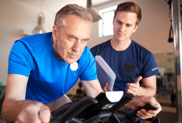 Senior Man Exercising On Cycling Machine Being Encouraged By Personal Trainer In Gym