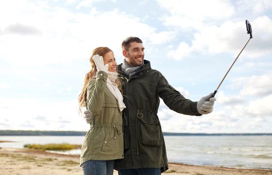 technology and people concept - smiling couple taking picture by smartphone on selfie stick on beach in autumn