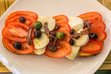 Lunchtime snack of tomatoes, cheese. anchovies and olives on bread, served on a white plate.