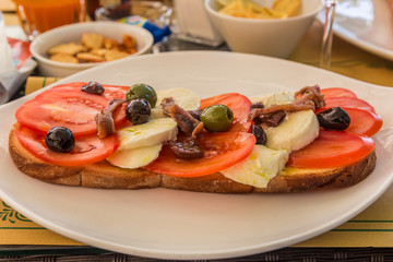 Lunchtime snack of tomatoes, cheese. anchovies and olives on bread, served on a white plate.