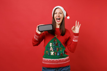 Merry young Santa girl keeping mouth wide open, showing OK gesture, holding portable wireless...