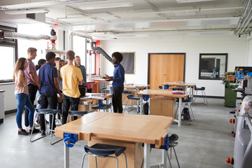 Group Of High School Students Standing Around Work Bench Listening To Teacher In Design And Technology Lesson