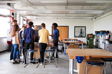 Group Of High School Students Standing Around Work Bench Listening To Teacher In Design And Technology Lesson