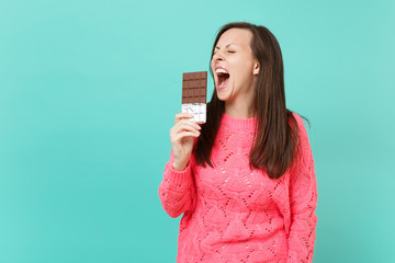 Crazy young girl in knitted pink sweater keeping eyes closed hold in hand biting chocolate bar isolated on blue turquoise wall background studio portrait. People lifestyle concept. Mock up copy space.