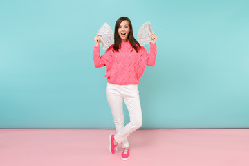 Full length portrait excited woman in knitted rose sweater, white pants holding lots dollars banknotes isolated on bright pink blue pastel wall background in studio. Money concept. Mock up copy space.