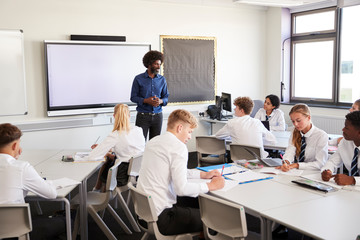 Male High School Teacher Standing Next To Interactive Whiteboard And Teaching Lesson To Pupils Wearing Uniform - obrazy, fototapety, plakaty