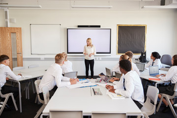 Female High School Teacher Standing Next To Interactive Whiteboard And Teaching Lesson To Pupils...
