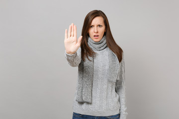 Distempered young woman in gray sweater, scarf showing stop gesture with palm isolated on grey background. Healthy fashion lifestyle, people sincere emotions, cold season concept. Mock up copy space.