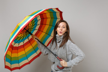 Amazed young woman in gray sweater, scarf holding colorful umbrella isolated on grey background in...