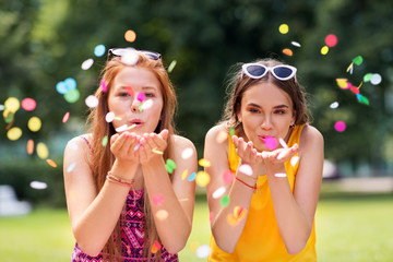 people and leisure concept - happy teenage girls or friends blowing confetti off hands at summer park