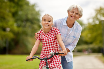 family, leisure and people concept - happy grandmother teaching granddaughter to ride bicycle at summer park