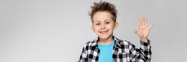 A handsome boy in a plaid shirt, blue shirt and jeans stands on a gray background. The boy smiles and shows the palm © Ivan Traimak