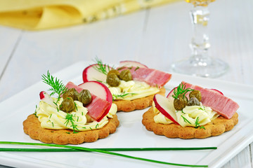 Canapes with goat cheese, red radish, egg slice and capers  and dill on white platter