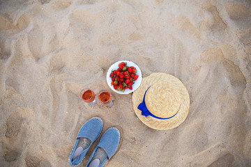 Fototapeta na wymiar a straw hat, two glasses of wine and a plate of strawberries on a sandy beach.