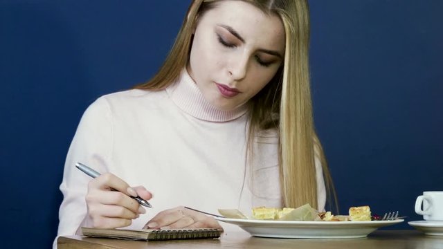 Young blonde girl eating waffles for breakfast, drinking tea and taking notes in notepad. 4K