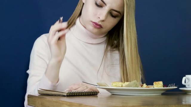 Young blonde girl eating waffles for breakfast, drinking tea and taking notes in notepad. 4K