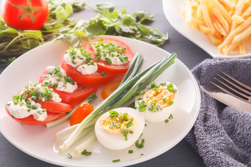 Half boiled eggs with tomatoes, green onions sprinkled with greens in a white plate on a gray wooden table. Close-up