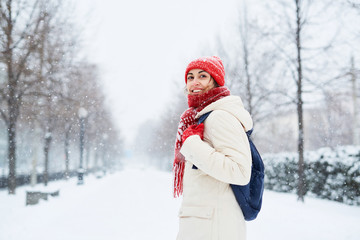 Cheerful smiling woman in white down jacket and red cap, scarf and mittens walking on the snowy street after blizzard in city. winter city after blizzards and snowfall.