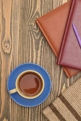 Blue tea cup with notebooks, a pen and a scarf on wooden table. Vertically.
