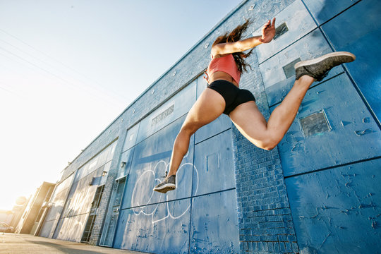 Low angle view of female athlete running along sidewalk past blue building covered in graffiti.