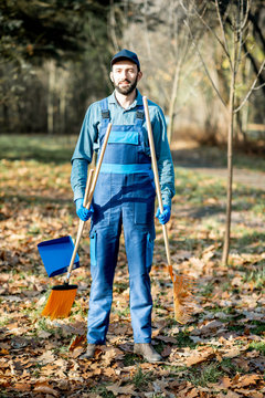 Portrait of a professional male sweeper in blue uniform with sweeping tools in the garden during the autumn time