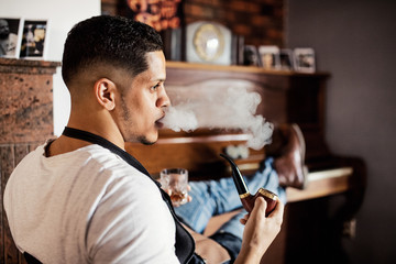 Young hispanic haidresser and hairstylist sitting in barber shop, smoking a pipe.