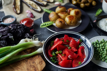 Delicious dinner table for two with fried potatoes, organic tomatoes salad, olives, green onion, eggplants on dark stone background.