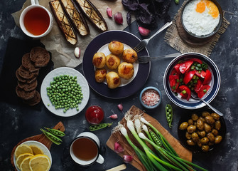 Flat lay of delicious dinner table for two with fried potatoes, organic tomatoes salad, olives, green onion, eggplants, eggs on dark stone background.