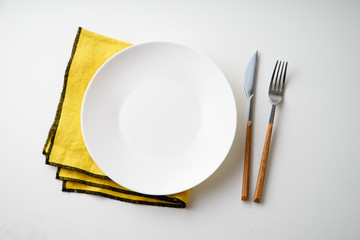 White empty plate with knife and fork and yellow textile napkin on white background