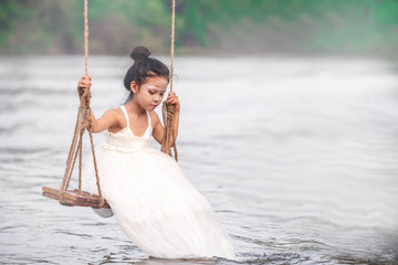 Little kid lonely playing swing along the riverside. Young girls in white dress as angle playing alone in the countryside.