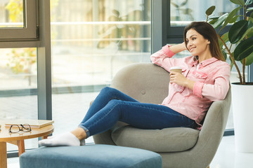 Young smiling woman sitting on sofa while drinking hot coffee. Young brunette woman thinking at home in a leisure time. Happy girl relaxing at home on big chair.