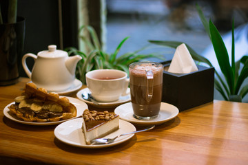 Sweet breakfast made of cacao drink , cheescake , wafer with chocolate and banana , teapot on wooden table. White simple dinnerware and green plants on background with window.