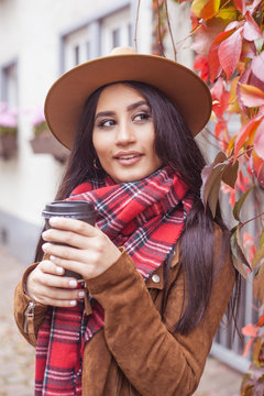 Young beautiful Turkish woman walks in a European city. A girl holds a glass of coffee. Happy brunette in a hat. Autumn. Fashion. Winter.