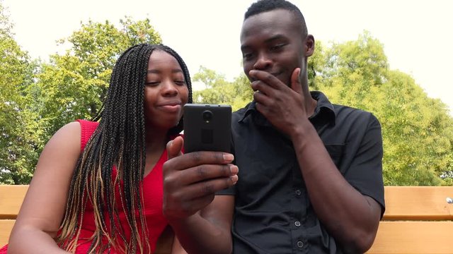 A black man and a black woman sit on a bench in a park, look at a smartphone and laugh - closeup from below