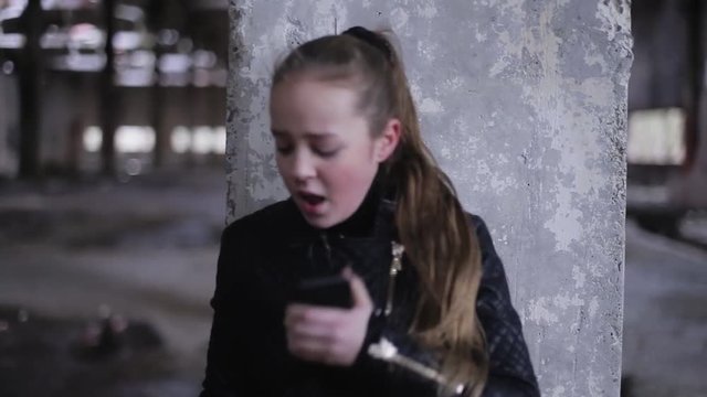 Little Caucasian girl in black clothes running from killer. Teen with gun in hand hiding, calling for help. Prosecution. Outdoors. Gray colors. Abandoned factory.