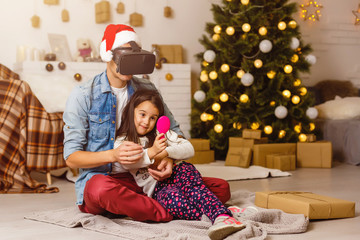 Dad and daughter using VR headset while sitting next to Christmas tree.