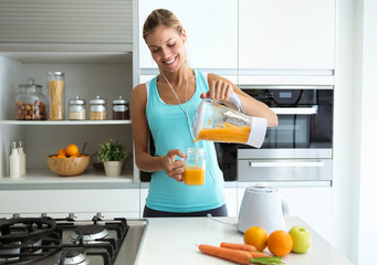 Beautiful young sporty woman serving detox orange juice into glass while listening music in the...